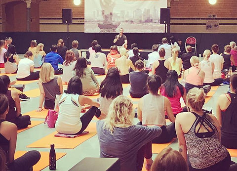 An image of Christoph sitting down on a riser stage, talking to a large group of people who are sitting down on yoga mats. 