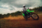 Motorcyclist on green bike kicking up dust on a dirt trail under cloudy skies.