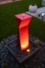 Red geometric fountain with water flowing over it, on black pebbles. Illuminated in a garden setting with green grass and a white planter.