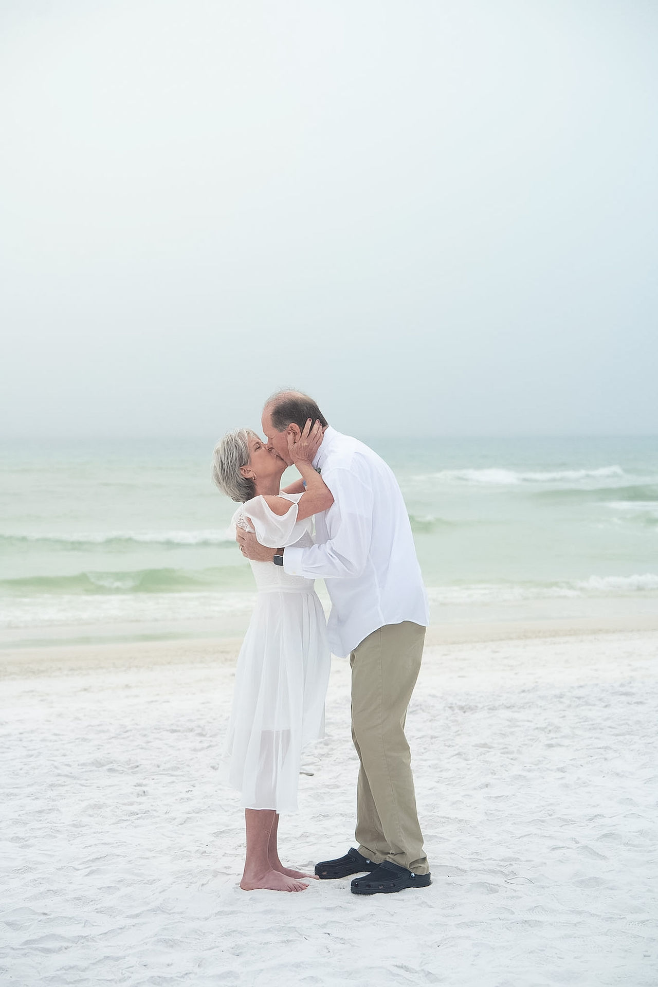 Couple kissing on the beach dressed in white for their wedding day.