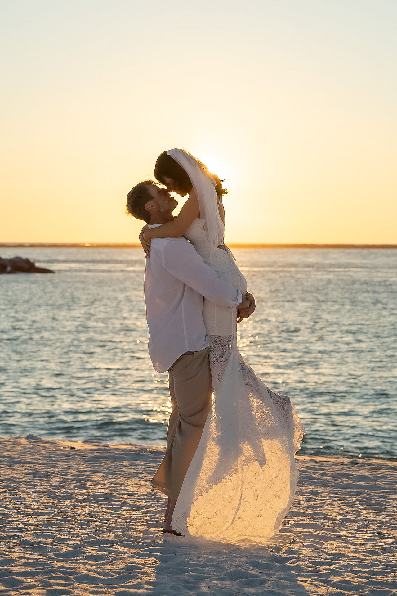 Groom picking up his bride and holding her in front of beach sunset.