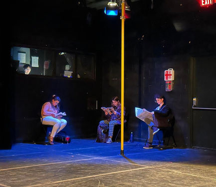 photo of stage, with a yellow subway pole and three actors, sitting and reading different books. Behind the actors and to the left, the stage manager/ subway conductor is seen behind glass