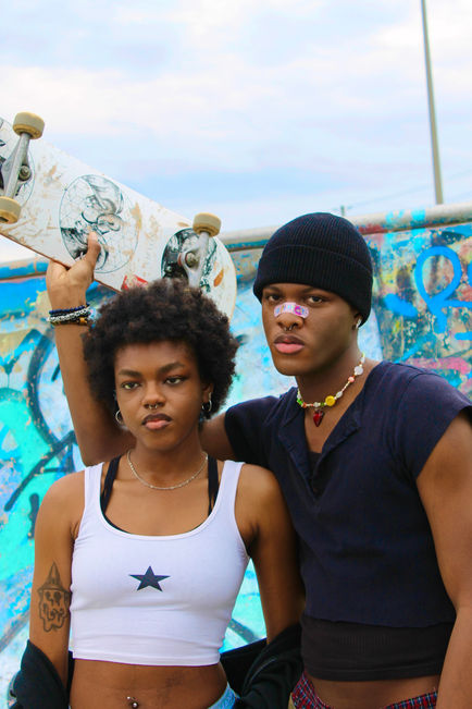 Two people posed in a skate park. The one on the left has dark skin and is wearing baggy alternative clothing. The one on the right has dark skin and is holding up a skateboard