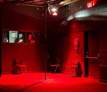 Empty stage with three chairs and a subway pole, lit red, with the stage manager in the background in a booth behind glass
