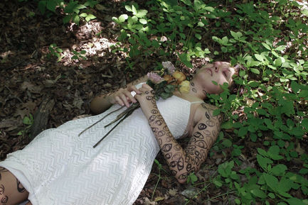 A photo of a person with makeup to look like they're decaying and illustrations of eyes covering them laying on a forest floor with flowers on their chest