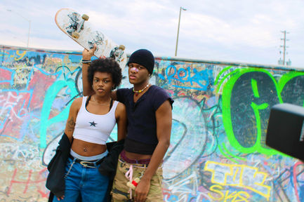 Two people posed in a skate park. The one on the left has dark skin and is wearing baggy alternative clothing. The one on the right has dark skin and is holding up a skateboard