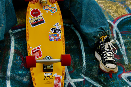 A yellow skateboard with colourful stickers leaned against someones leg