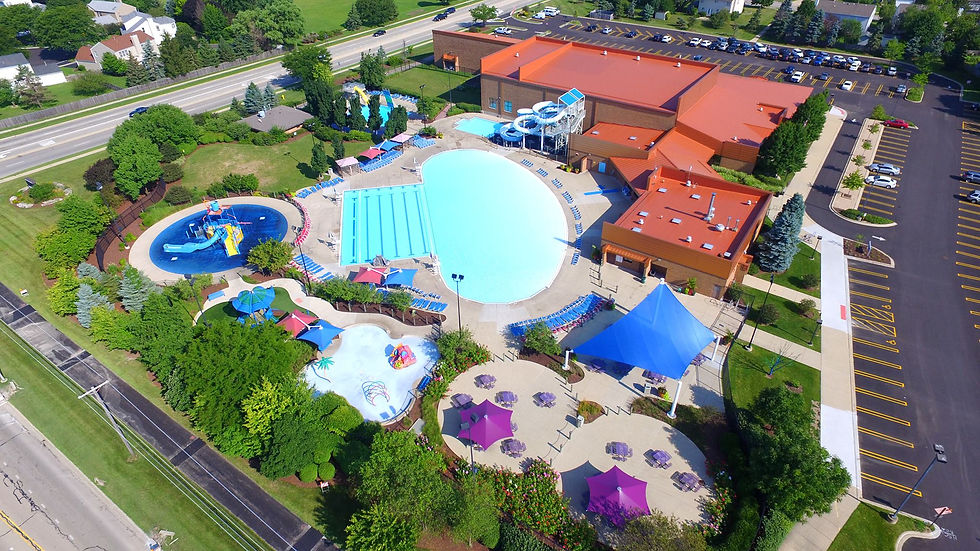 Outdoor Arial photo of Coral Cove Water Park featuring a lap pool, zero depth entry pool, water slides, table areas, water playground and splash pad area, orange building and more.