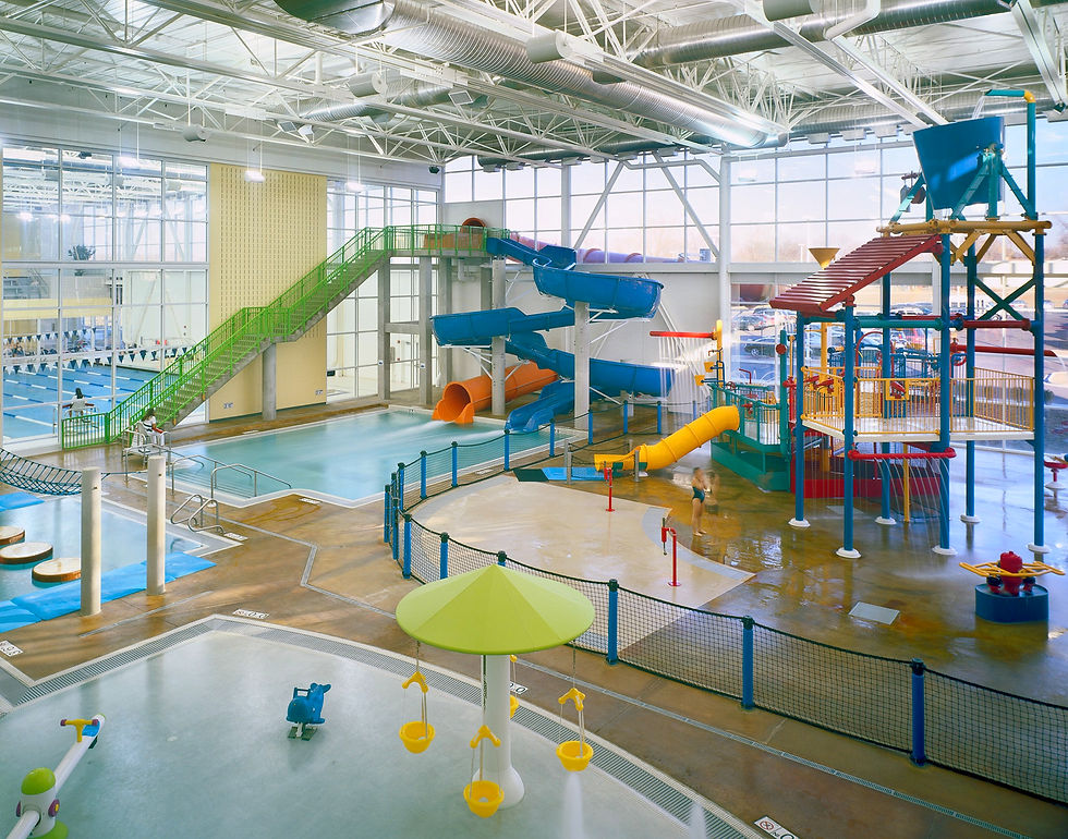 photo of Vaughan Aquatic Center inside water park, featuring water playground, and splash pad for younger kids, green stair case leading to a blue and a purple water slide, and lap pool in the distance.