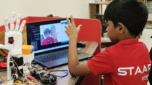 Young boy interacts with a robotic hand via laptop. The screen shows his mirrored actions. Tech equipment on wooden table.