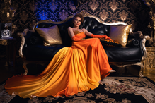 A woman in an orange gown sits on a ornate black couch, gold pillows beside her. Luxurious, patterned wallpaper in the background.