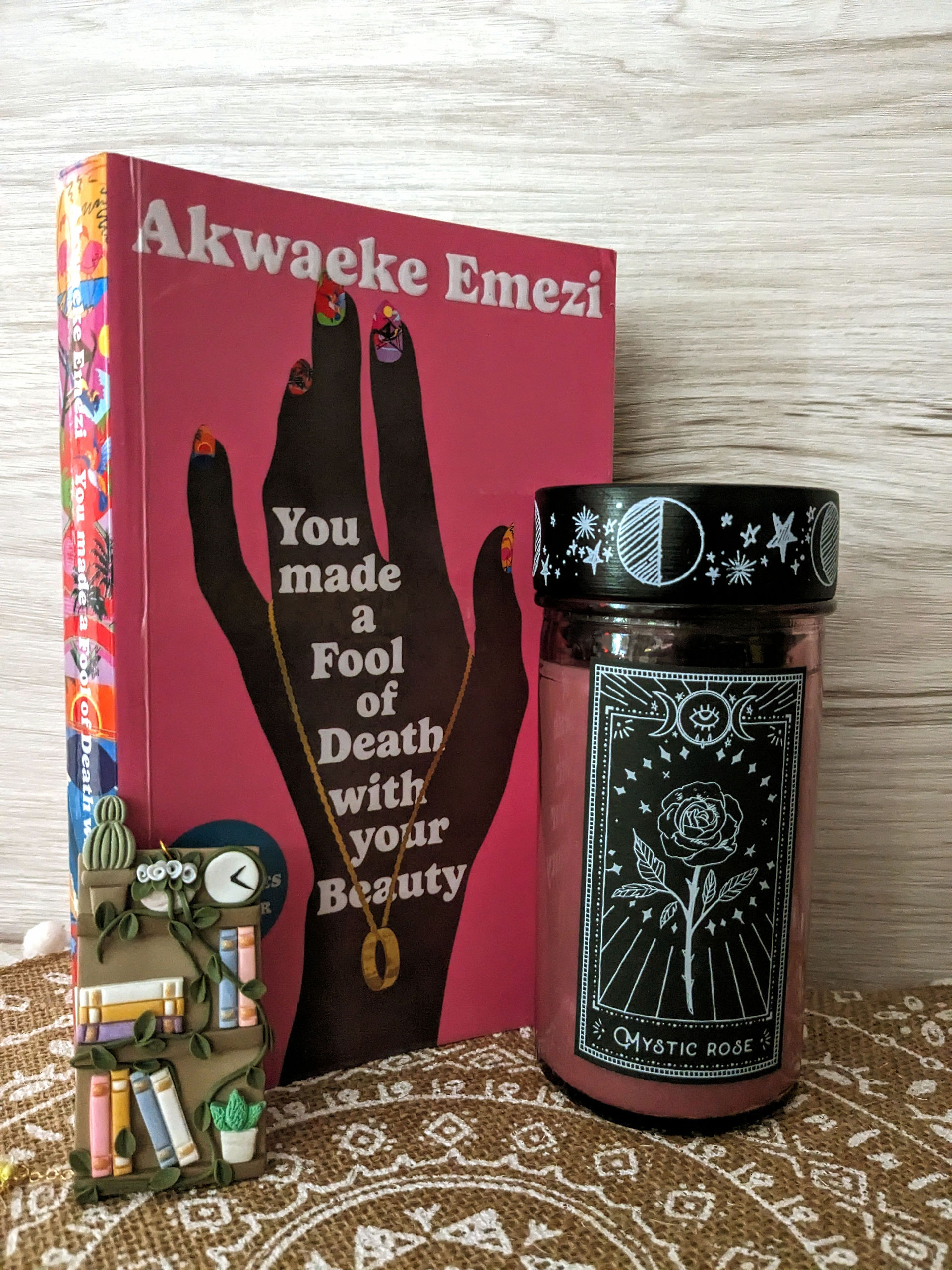 A posed photo from Rose Thomson's Instagram feed of the book You Made a Fool of Death With Your Beauty posed standing upright with a mini clay, 3D handmade bookshelf  and a candle. The colours all tone in with the ink background of the book, with the candle being a dusty pink with black label and lid. 