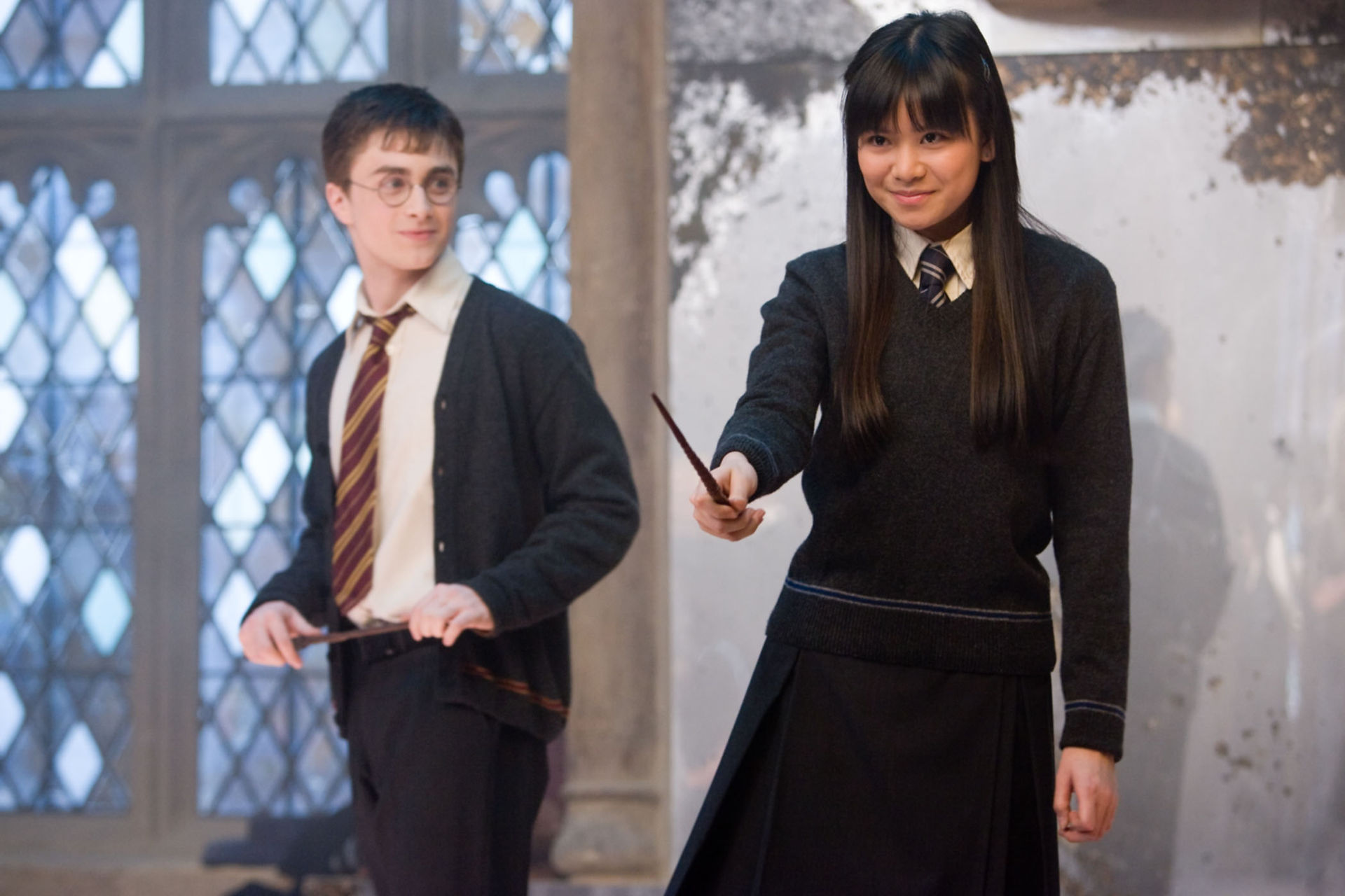 A screenshot from a Harry Potter movie, showing Harry Potter watching Cho Chang, who is the central figure in the shot. Cho and Harry both are in their Hogwarts uniforms and hold wands. They are in a classroom and Cho holds her wand out about to practice a spell. . 
