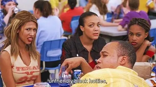 A screenshot from the movie Mean Girls which shows a cateteria scene, the focus of which is a group of Black or POC characters, and the subtitle shown reads "unfriendly black hotties", the stereotype assigned to their table clique in the movie. 