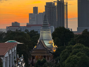 Sunset view of Wat Phnom temple surrounded by lush greenery, with modern Phnom Penh skyscrapers in the background, captured from TRIBE Phnom Penh Post Office Square hotel rooftop