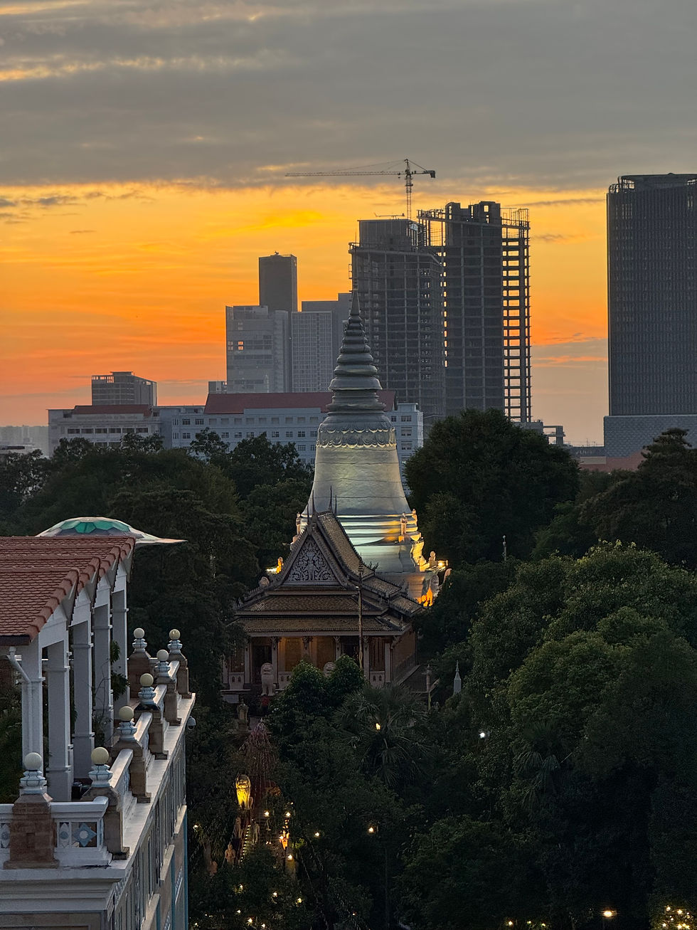 Sunset view of Wat Phnom temple surrounded by lush greenery, with modern Phnom Penh skyscrapers in the background, captured from TRIBE Phnom Penh Post Office Square hotel rooftop
