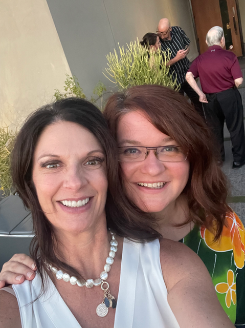 Two smiling women pose for selfie outdoors, with pearl necklace.