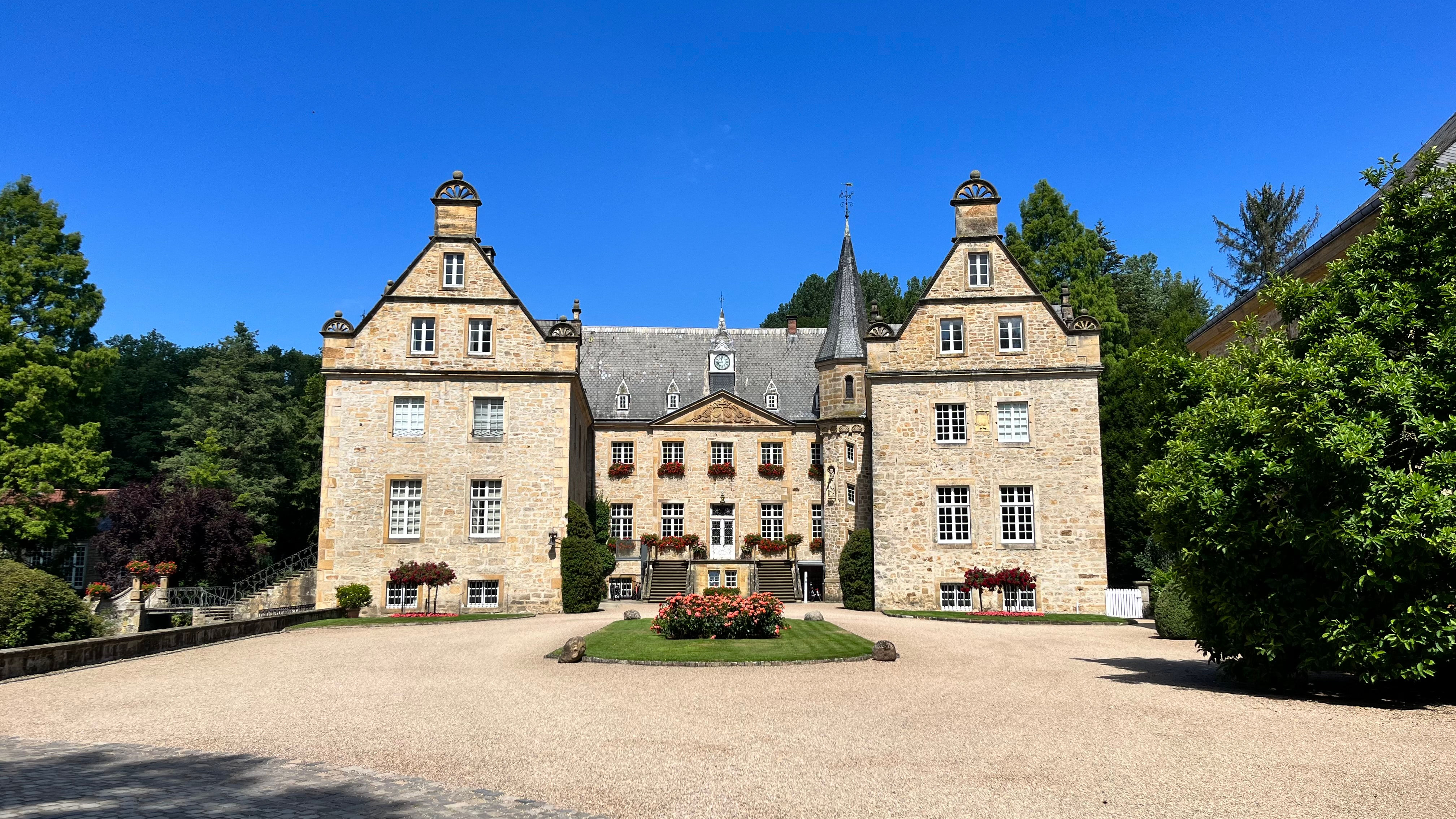 Ein doppelflügliges Schloss mit einem großen Vorplatz. Der Himmel ist strahlend blau