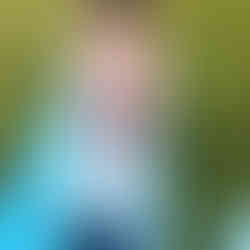 Woman sitting on a blue mat in a park, holding a baby. She smiles down at the baby, surrounded by greenery and parked cars.