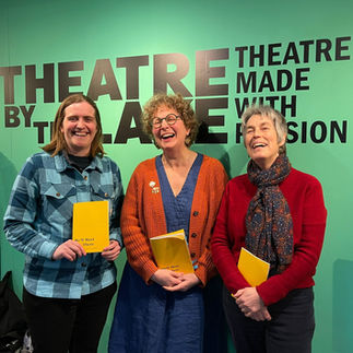 Three white women - me (Hilary), Alice and Ilse with our yellow books We'll Meet You There in front of a very bright teal wall that says Theatre By the Lake (which is in Keswick)