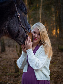 Schwangere Frau mit offenen blonden Haaren, weißer Strickjacke und lilanem Kleid schaut an die Nase ihres Pferdes geschmiegt in die Kamera