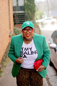 Stylish woman in green blazer with "In my healing era" t-shirt, with a red cross body, Green NY Yankees baseball cap,and leopard pants. 