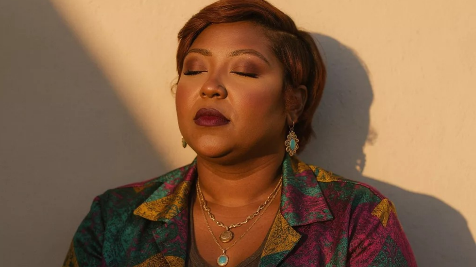 Woman with eyes closed in warm sunlight, wearing a colorful patterned jacket and turquoise jewelry against a plain wall.