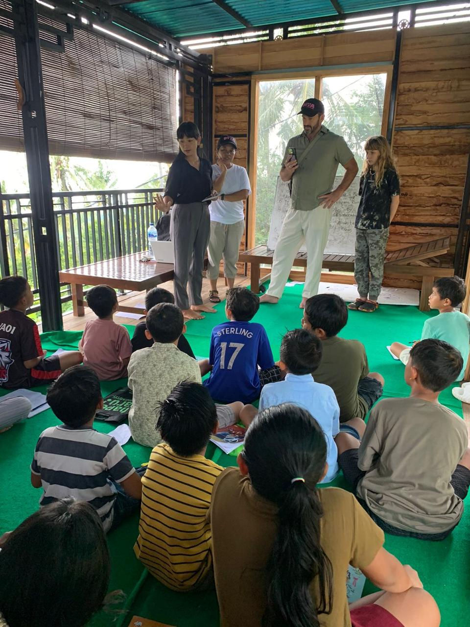 Adults present to seated children in a wooden room with green flooring. A man holds a phone, others have papers. Windows show greenery outside.