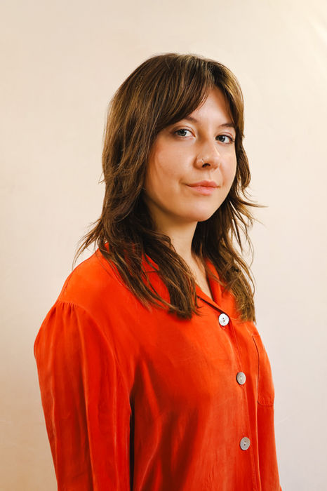 A relaxed portrait of a woman wearing an orange shirt, photographed in three-quarter profile against a neutral backdrop at a London portrait studio, suitable for creatives or personal branding.