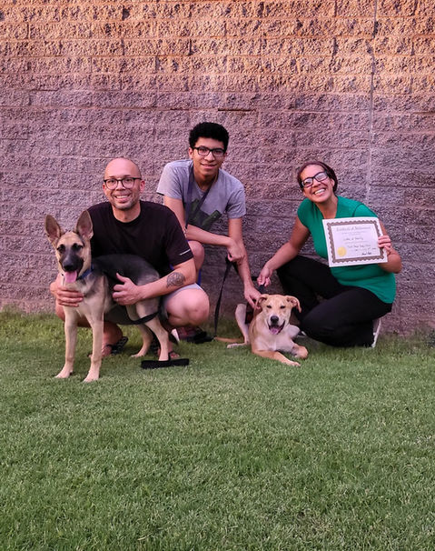 Three people kneeling on grass with two dogs in front of a brick wall; one person is holding a 6 Paws Training Camp certificate and smiling.