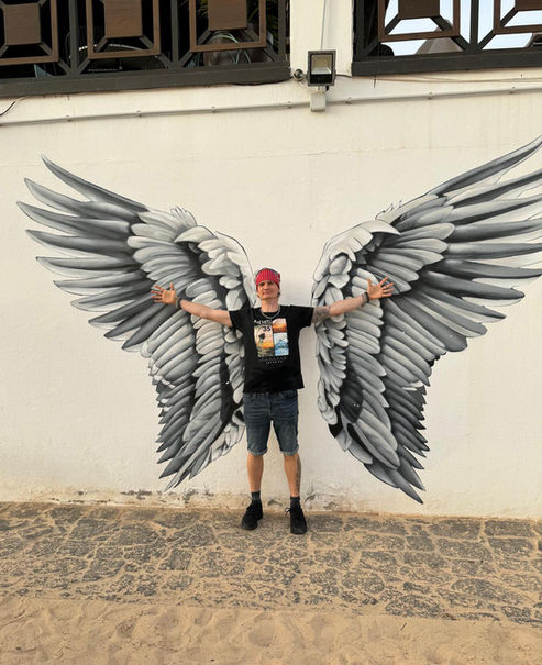 A man stands with his arms outstretched in front of a wall with angel wings painted on it, making it look as if he has wings.