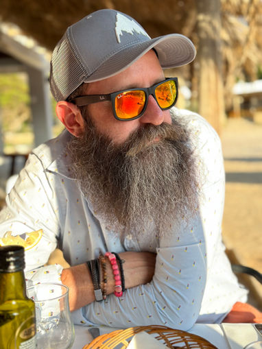 A person wearing a grey cap, sunglasses and a long beard sits relaxed at a table outdoors, the sunlight reflecting off their glasses and creating a warm, summery atmosphere.