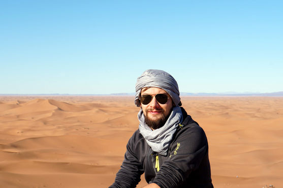 A person wearing sunglasses and a grey turban sitting in a vast desert landscape with sand dunes under a clear blue sky.