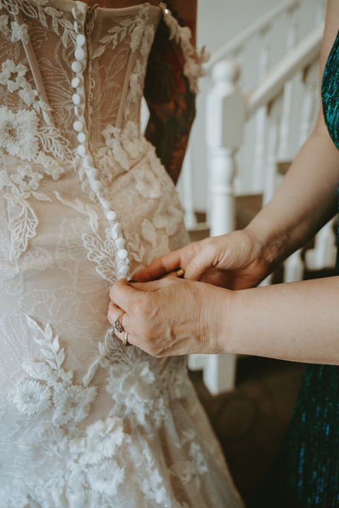 Bride’s mother helping her fasten wedding dress during morning preparations.