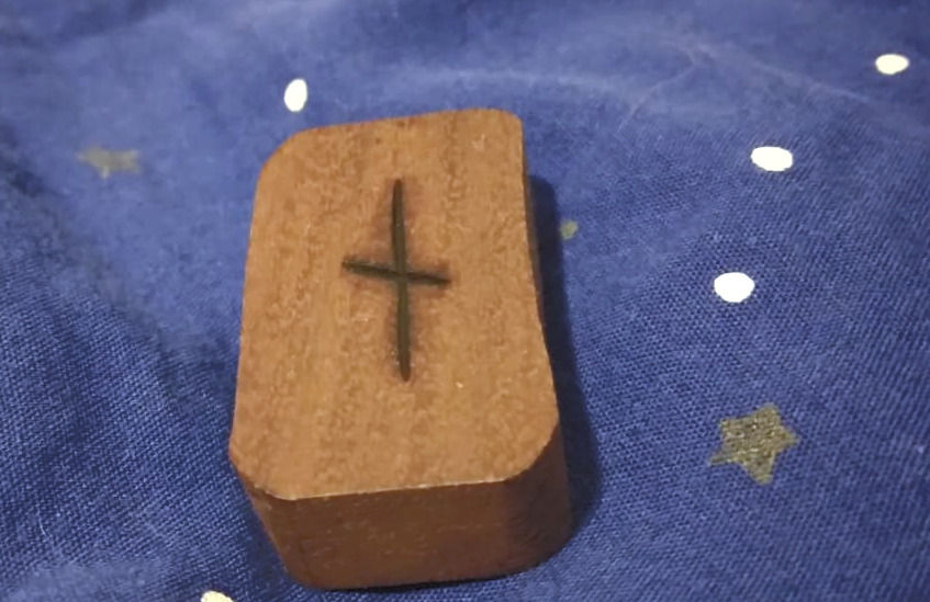 A brown wooden rectangle is carved with a Naudhiz rune, which is like a lower-case letter T