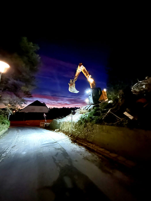 Beleuchteter Bagger bei Nacht auf einem Erdhaufen vor dämmerndem Abendhimmel.