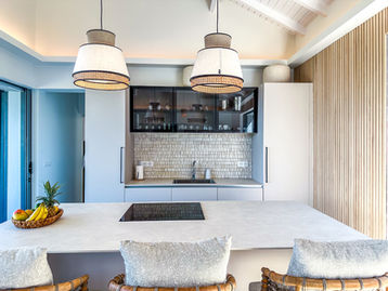 Designer kitchen with white central island, woven chairs, natural fiber pendant lights and textured wall at Baie du Paradis