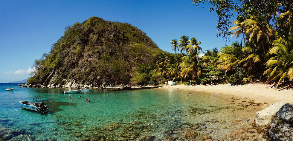 Vue panoramique de la plage du Pain de Sucre en Guadeloupe, avec une eau cristalline aux nuances turquoise et bleu profond. Le sable est doré et fin, bordé par une végétation tropicale luxuriante qui apporte de l'ombre naturelle. À l'arrière-plan, le célèbre mont Pain de Sucre s'élève majestueusement, ajoutant une touche spectaculaire au paysage côtier. Quelques bateaux sont amarrés au large, créant une scène de sérénité et d’évasion dans ce coin de paradis caribéen.