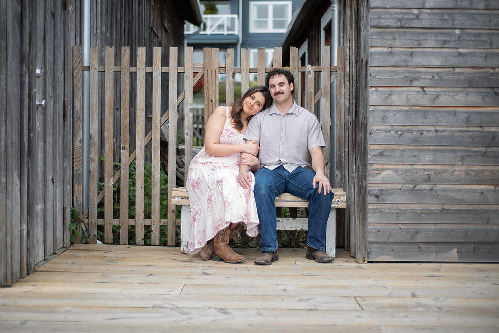 Engaged couple sitting on a wood bench on the boardwalk at the Shipyards in Richmond, with wood houses to either side of them. There is a nice colour palette with the pale of the weathered wood, her light pink floral dress and cowgirl boots, and his grey button down, jeans and cowboy boots.