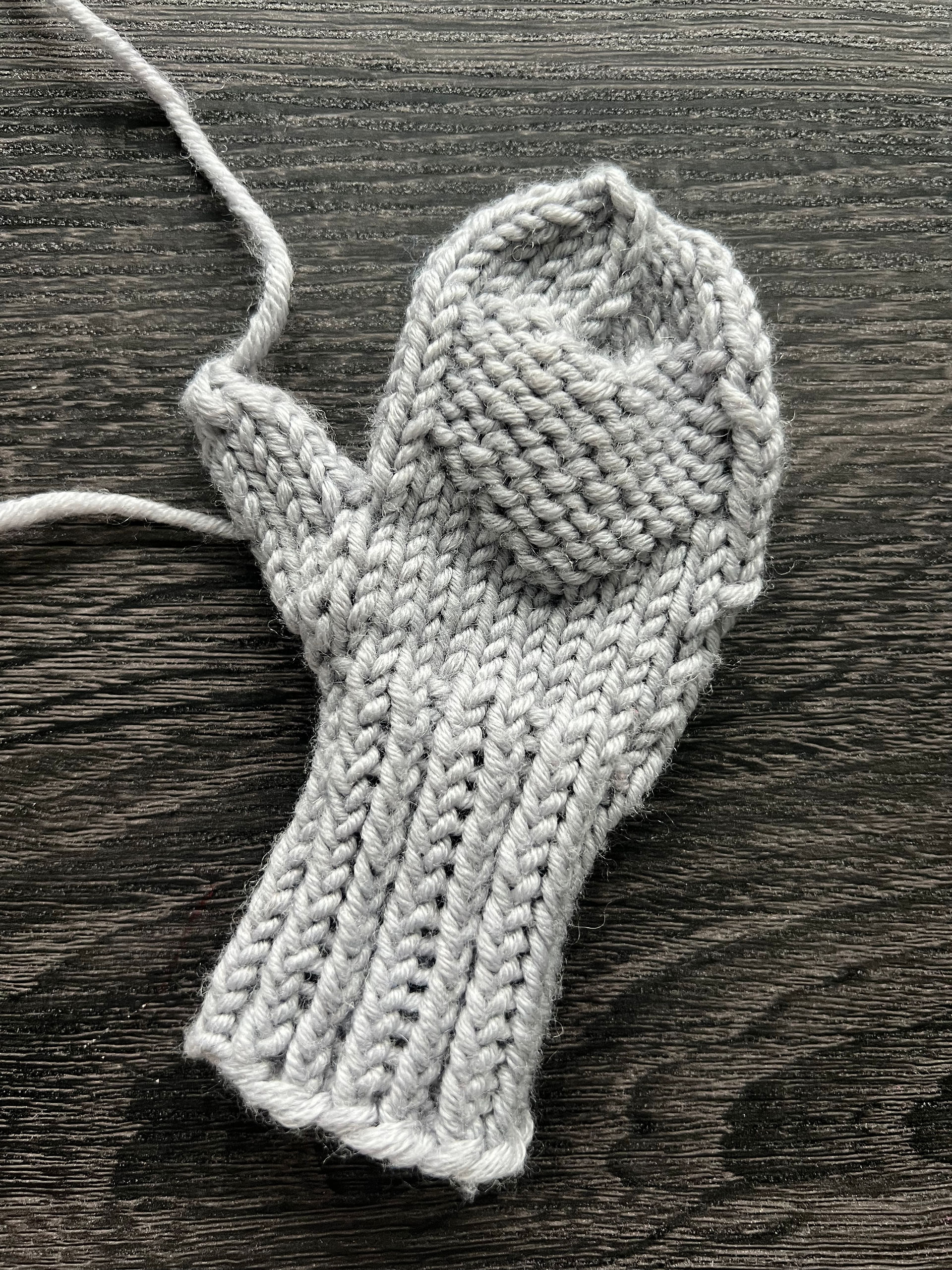 A single mitten lays on table with ends hanging from thumbs. Simple knit with gray yarn and a purled heart on the palm.