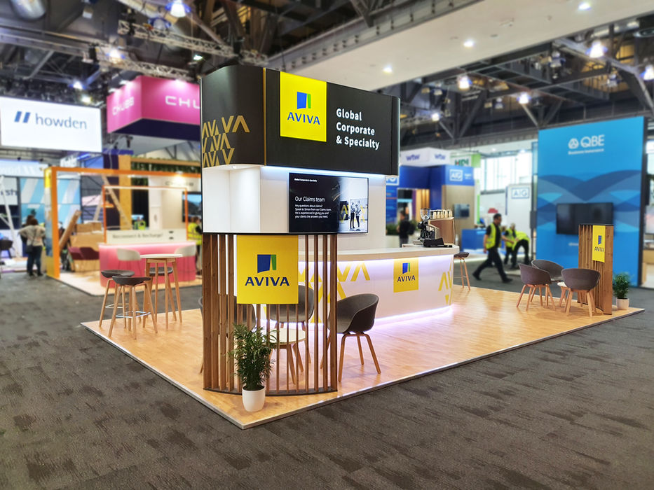 Exhibition stand for Aviva at the Airmic Conference. Branded graphics, a digital display, wraparound counter, coffee bar and meeting area make up the stand space.