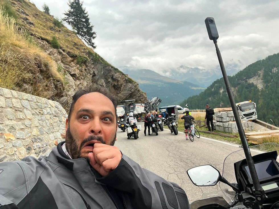 Shocked man takes a selfie on a mountain road with motorcycles, cyclists and trucks under cloudy alpine peaks.