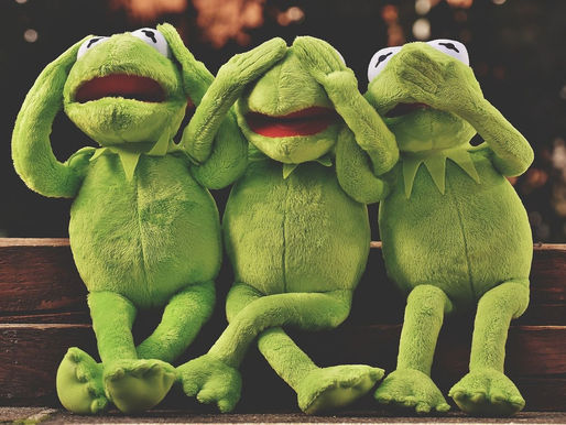 Three green Kermit-style puppets sit on a bench, mimicking see/hear/speak no evil with playful, amused expressions.