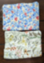 Flatlay of 2 folded squares of fabric vertically shown. The top is a light and cerulean blue floral with red and white florals scattered across it. Background is white. The bottom fabric is white with long stemmed flowers that look like daisies, mums, and yellow wildflowers.