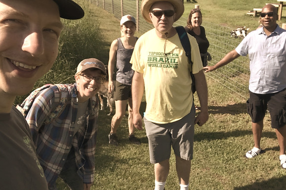 6 people, a mix of ages and abilities, posing for a selfie in a green space. Goats in the background.