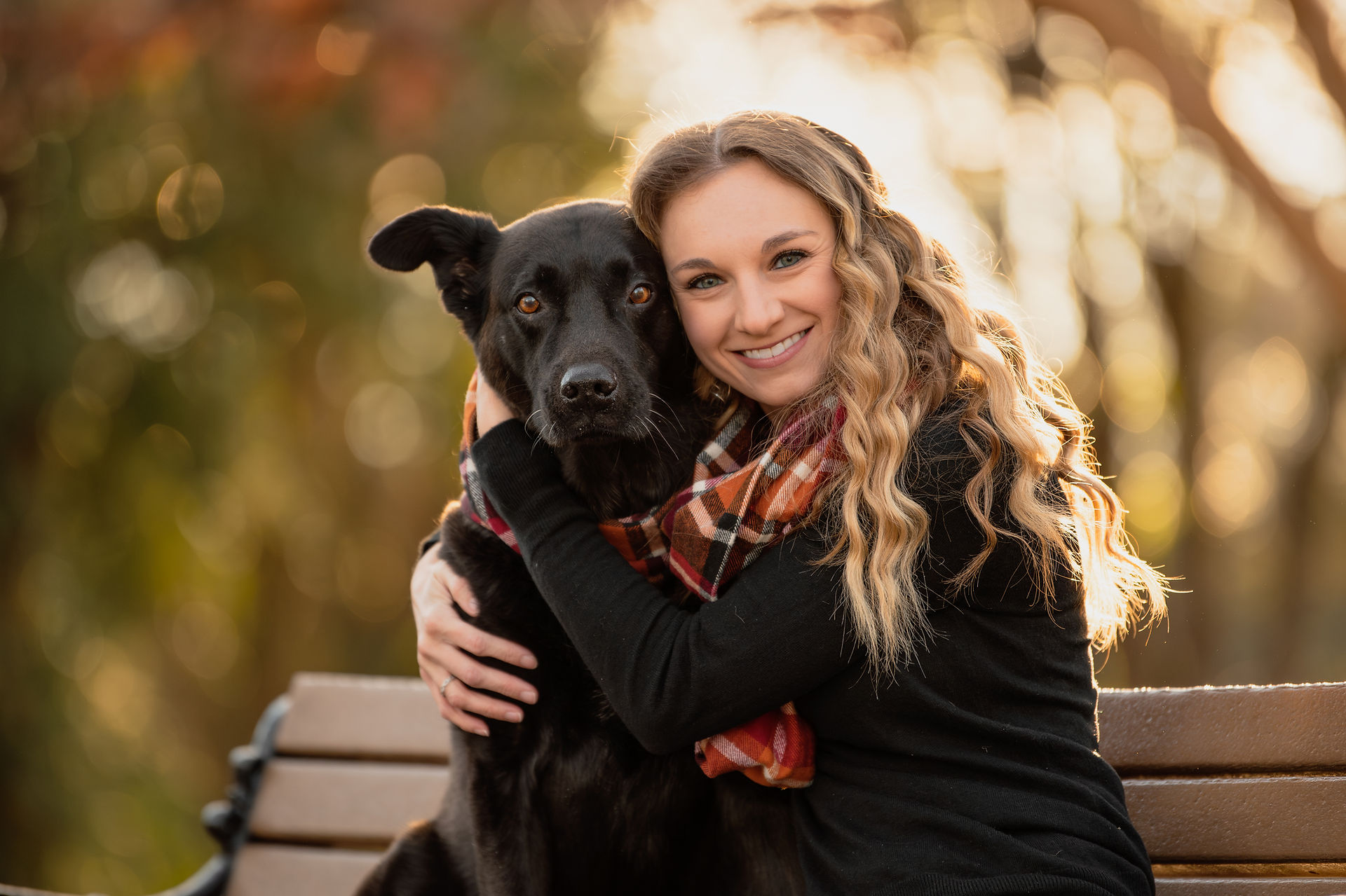 A girl with curly hair sitting on a bench with her black dog. The dog has semi-pointy airplane ears and they are both looking directly at the camera.