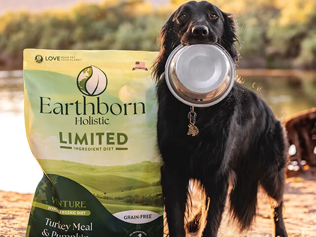 Dog holding silver bowl while standing beside a bag of earthborn unlimited dog food