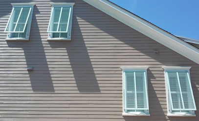 Light green decorative Bahama shutters with aluminum inside mounted adjustable support arms installed on the windows of a weathered gray coastal home with white trim.