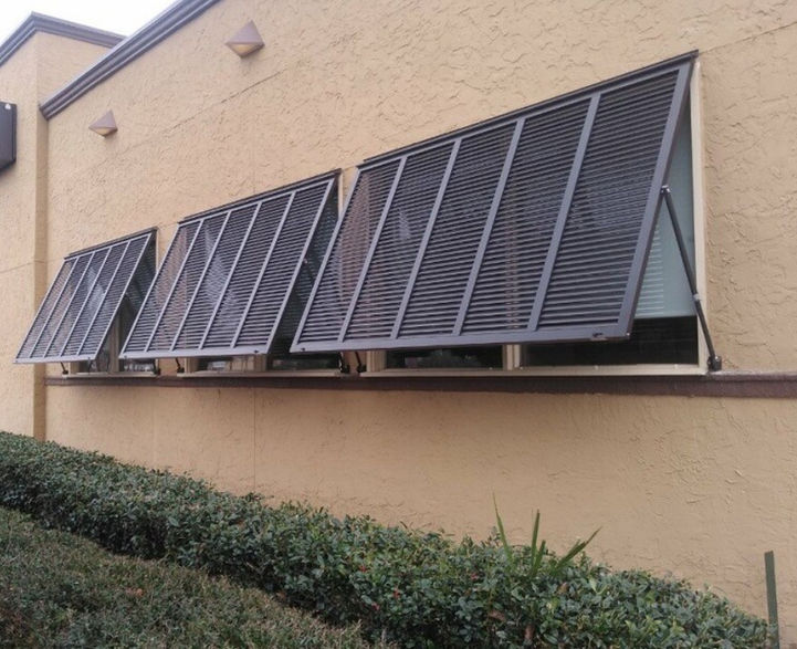 Chocolate brown, impact Bahama shutters with matching outside mounted adjustable support arms installed on a tan brick commercial property with cream trim.