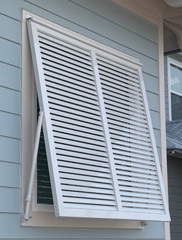 Gloss white decorative Bahama shutters with matching outside mounted adjustable support arms installed on a first floor window of a light aqua home with white trim.